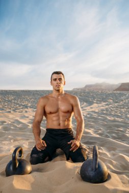 Erkek sporcu, güneşli bir günde çölde idmandan sonra kumda oturuyor. Sporda güçlü motivasyon, açık hava eğitiminde güçlü.