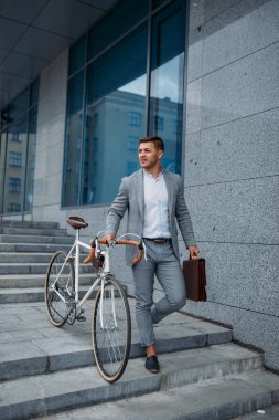 Bisikletli bir iş adamı şehir merkezindeki ofis binasının merdivenlerinden aşağı iniyor. Şehir caddesinde çevre taşımacılığı yapan iş adamı, şehir tarzı.