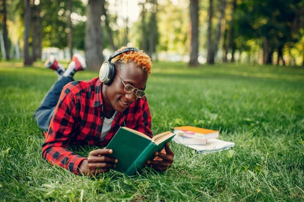 Kulaklıklı Afrikalı Bir Öğrenci Çimlerin Üzerinde Uzanıyor Yaz Parkında Kitap — Stok fotoğraf