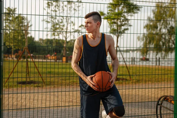 Male basketball player with ball standing at the mesh fence on outdoor court, side view. Male athlete in sportswear on streetball training, summer stadium