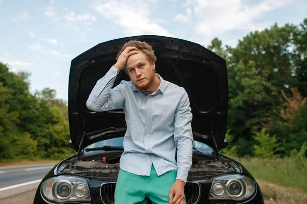 Tired young man at the opened hood, car breakdown. Broken automobile or repairing of vehicle, trouble with auto engine on highway