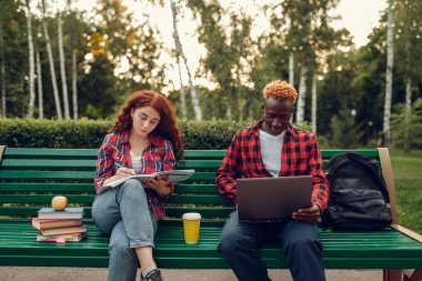 Yaz parkında bankta oturan iki öğrenci. Erkek ve dişi beyaz gençler açık havada okuyor.