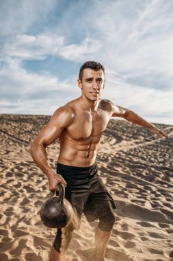 Erkek sporcu, güneşli bir günde çölde kettlebell ile egzersiz yapıyor. Uçan kum etkisi. Sporda güçlü motivasyon, açık hava eğitiminde güçlü.