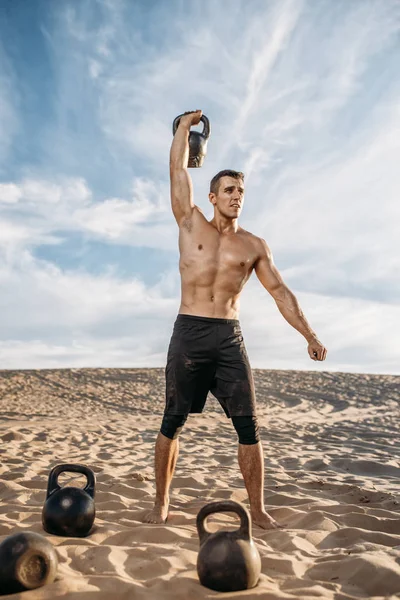 Muscular male athlete doing exercises with kettlebell in desert at sunny day. Strong motivation in sport, strength outdoor training, sportsman with weights