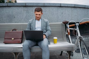Bisikletli bir iş adamı şehir merkezindeki ofis binasının bankında kahve içer. Şehir caddesinde çevre taşımacılığı yapan iş adamı, şehir tarzı.