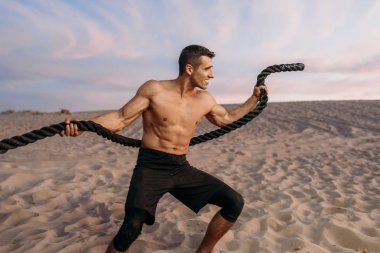 Kaslı adam güneşli bir günde çölde iple egzersiz yapıyor. Sporda güçlü motivasyon, açık hava eğitiminde güçlü.