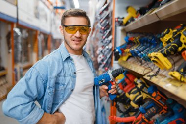 Hırdavatçıda elinde elektrikli tornavida tutan bir erkek alıcı. Müşteri dükkandaki mallara bakar, süpermarketten alışveriş yapar.