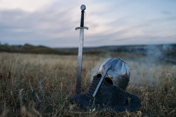 Sword stuck in the ground and metal knight helmet, nobody, great warriors symbol in the field