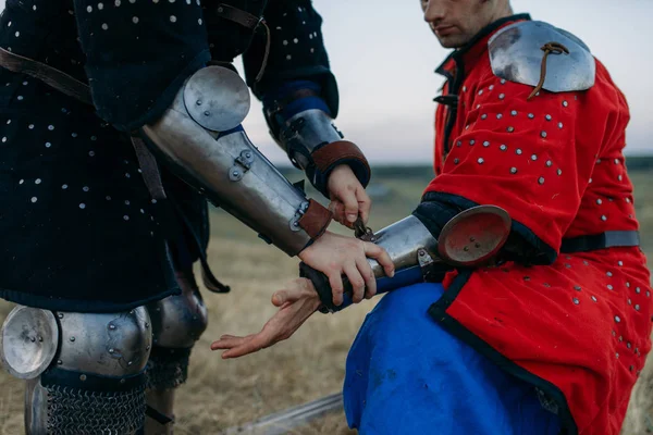 Ortaçağ şövalyesi arkadaşının zırh giymesine yardım ediyor, büyük bir turnuva. Zırhlı eski bir savaşçı tarlada poz veriyor.
