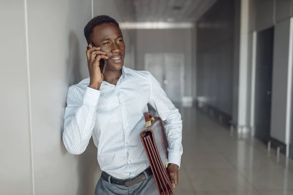 African businessman with briefcase talking by phone in office hallway