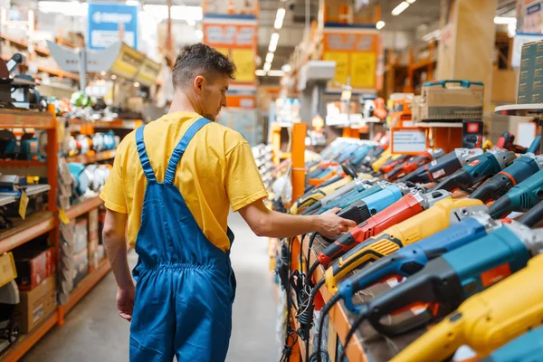 Male builder choosing angle grinder at the shelf in hardware store. Constructor in uniform look at the goods in diy shop