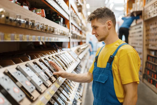 Male builder choosing door lock at the shelf in hardware store. Constructor in uniform look at the goods in diy shop