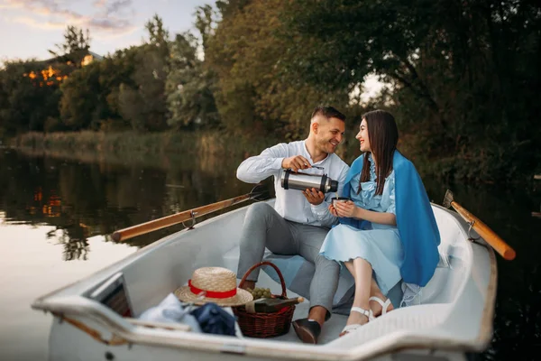 Love couple with thermos in a boat on quiet lake at sunset. Romantic meeting, boating trip, man and woman walking along the river