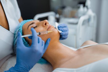 Eldivenli kozmetik uzmanı tedavi masasındaki kadın hastaya çene botoksu veriyor. Güzellik salonunda gençleştirme prosedürü. Şırıngalı ve kadınlı doktor, kırışıklıklara karşı estetik ameliyat.