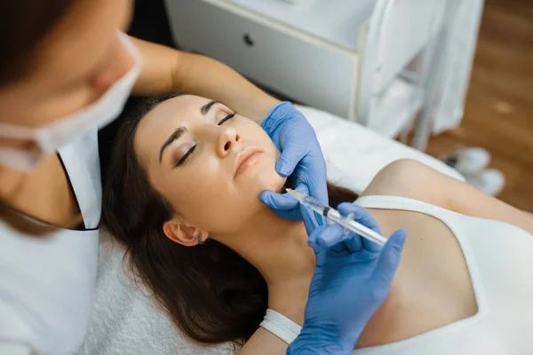 Cosmetician in gloves gives chin botox injection to female patient on treatment table. Rejuvenation procedure in beautician salon. Doctor with syringe and woman, cosmetic surgery against wrinkles