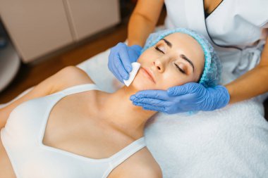 Kozmetik uzmanı yüz derisini kadın hastaya temizliyor. Güzellik salonunda gençleştirme prosedürü. Doktor ve kadın, kırışıklıklara ve yaşlanmaya karşı estetik ameliyat.
