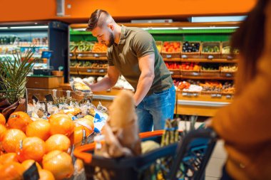 Markette alışveriş arabası olan çift. Erkek ve kadın marketten meyve ve sebze alıyor, müşteriler alışveriş yapıyor.
