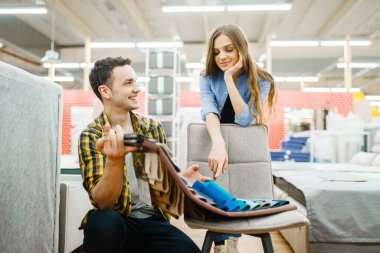 Genç çift mobilya mağazasında yatak süsü seçiyor. Kadın ve erkek dükkanda yatak odası örnekleri arıyor, karı ve koca modern iç mekanlar için ürünler satın alıyor.
