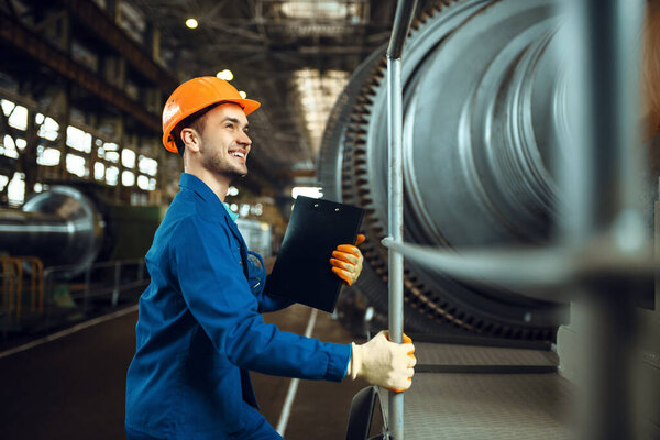 Male worker with notebook, large turbine on background, plant. Industrial production, metalwork engineering, machines manufacturing