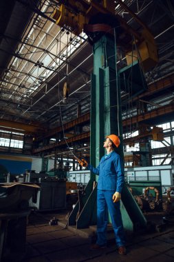 Üniformalı ve kasklı bir erkek işçi fabrikada vinç kullanıyor. Endüstriyel üretim, metal mühendisliği, güç makineleri üretimi