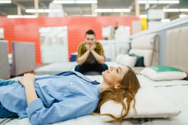 Genç mutlu çift mobilya mağazasından yatak alıyor. Kadın ve erkek dükkanda yatak odası örnekleri arıyor, karı ve koca modern iç mekanlar için ürünler satın alıyor.