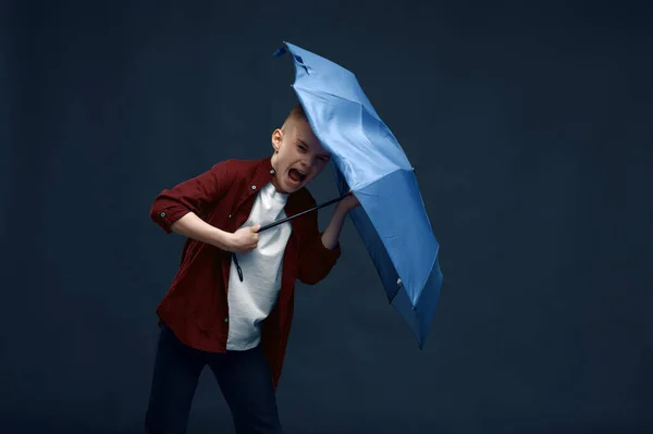 Little boy with a scared face holds an umbrella in studio, wind effect, powerful airflow. Children with developing hairs, kids isolated on dark background, child emotion
