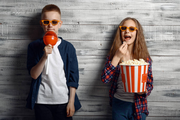 Little boy and girl in sunglasses holds popcorn and big lollipop in studio. Children and sweets, kids isolated on wooden background, child photo session