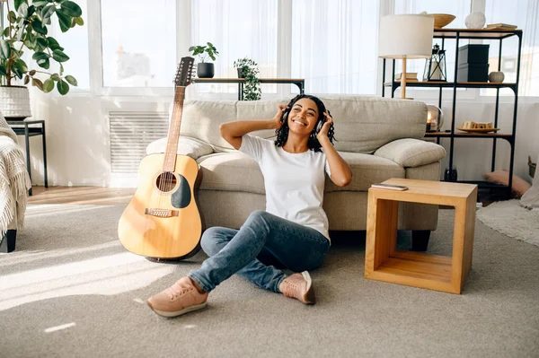 在家里用耳机微笑的女人 背景上的吉他 美丽的女士带着乐器在房间里放松 女性音乐爱好者休息 — 图库照片