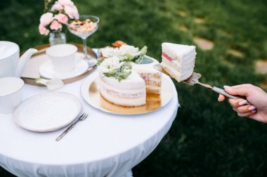 Masa düzenleme, çay partisi, kadın eli pastayı yakın plan çeker, yan görüş. Beyaz masa örtüsü üzerinde lüks gümüş takımlar, açık havada sofra takımı.