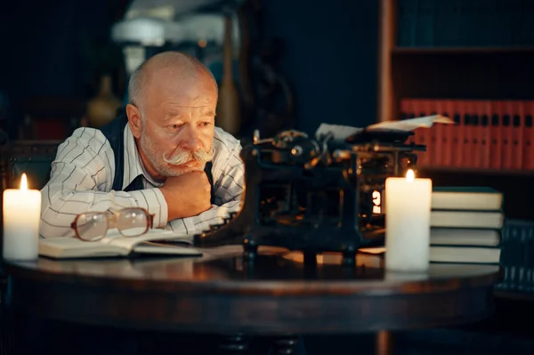 Adult writer works on vintage typewriter with candle light in home office. Old man in glasses writes literature novel in room with smoke, inspiration