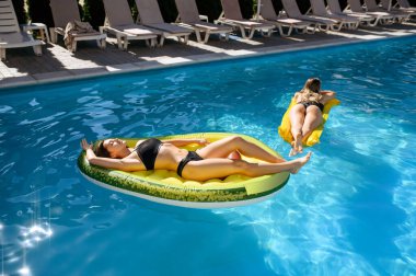 İki seksi kadın tatil köyündeki havuzda şişme yatakta güneşleniyor. Güzel kızlar güneşli bir günde havuz başında dinlenirler, yaz tatillerinde çekici kadınlar olur.