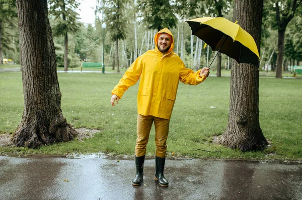 風の強い雨の日に夏の公園で傘を歩く男 雨のケープとゴム製のブーツを着た男性 路地の雨の天気 — ストック写真