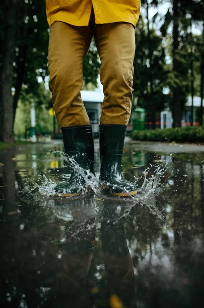 雨のケープとゴムブーツの男性は水溜りでジャンプし 路地に雨の天気 男は夏の公園 雨の日にポーズ 水の保護 — ストック写真