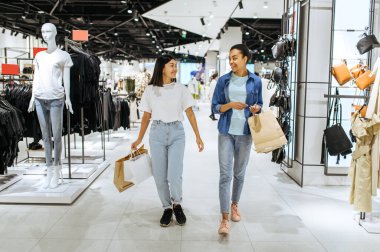 Karton çantalı gülümseyen kız arkadaşlar giyim mağazasında yürür. Moda mağazasında alışveriş yapan kadınlar, alışveriş meraklıları.