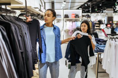 İki kız giyim mağazasında elbise asıyor. Moda mağazasında alışveriş yapan kadınlar, alışveriş kolikler, giysi arayan müşteriler.