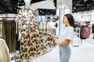 Giyim mağazasında elbise seçen neşeli bir kadın. Moda mağazasında alışveriş yapan kadın, alışverişçi, kıyafet arayan kadın.