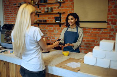 Kahvede erkek barista, siparişi bayan müşteri ödüyor. Adam kafeteryada taze espresso yapıyor, garson ve kadın barda tezgahta.