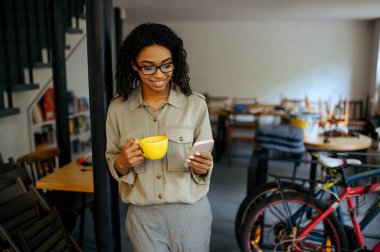Gözlüklü tatlı kız öğrenci kahve içiyor ve kafede telefon kullanıyor. Kadın, kahve, eğitim ve yemek konularında ders alıyor. Kampüs kafeteryasında okuyan kız.