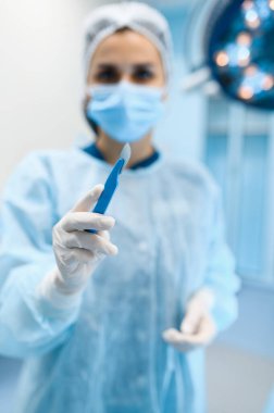 Önlüklü ve maskeli kadın cerrah ameliyathanede neşter arka planda lamba ve ameliyat olduğunu gösteriyor. Üniformalı doktor, klinik çalışanı, cerrahi tıp ve sağlık, hastanede sağlık hizmetleri.