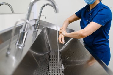 Maskeli kadın cerrah ellerini yıkıyor ve ameliyata hazırlanıyor. Üniformalı doktor, klinik çalışanı, ilaç ve sağlık, hastanede sağlık hizmetleri