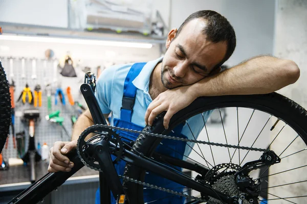 Bicycle repair in workshop, tired repairman sleeps at workplace. Mechanic in uniform fix problems with cycle, professional bike repairing service