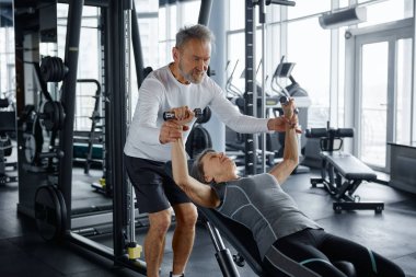 Yaşlı bir adam, bir spor salonundaki bankta oturan bir kadına halterleri kaldırırken yardım ediyor.