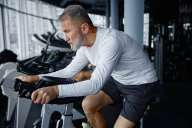 Spor salonunda egzersiz bisikletine binen bir adam diğer fitness meraklıları etrafını sarmışken antrenmanına odaklanıyor.