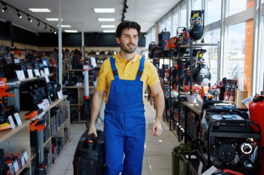 Bir işçi bir dükkândan geçerken elinde elektrikli süpürge taşır. Muhtemelen bir işe ya da işe hazırlanıyordur.