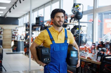 Mavi tulumlu bir adam bir mağazanın içinde kaynak makinesi tutuyor. İşine bakıyor ya da hazırlanıyor.