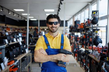 Güneş gözlüğü takan bir adam, elinde matkap varken bir mağazada duruyor ve satın alınabilecek aletleri gösteriyor.