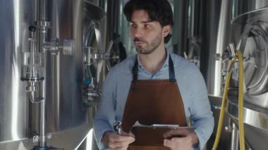 Bira fabrikasında çalışırken önlük giymiş bira üreticisi elinde bir pano tutuyor. Etrafı bira ekipmanları ve malzemelerle çevrili.