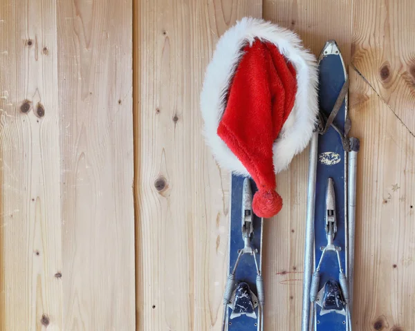 सांता क्लॉस की टोपी एक कुटीर के अंदर एक लकड़ी की दीवार के खिलाफ एक पुराने स्की से लटका हुआ — स्टॉक फ़ोटो, इमेज
