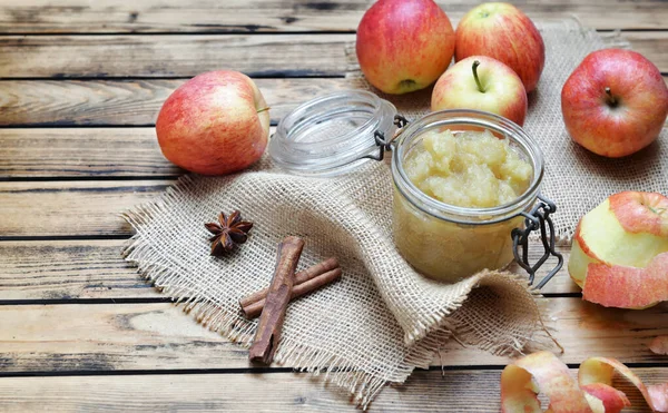 homemade apple sauce in a glass jar with red applesand stick of cinnamon on a wooden table
