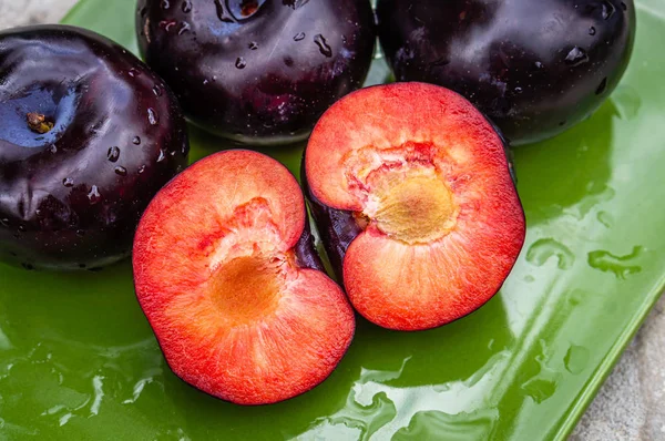 Tres frutos de ciruela morados oscuros y uno cortado en un plato verde —  Fotos de Stock